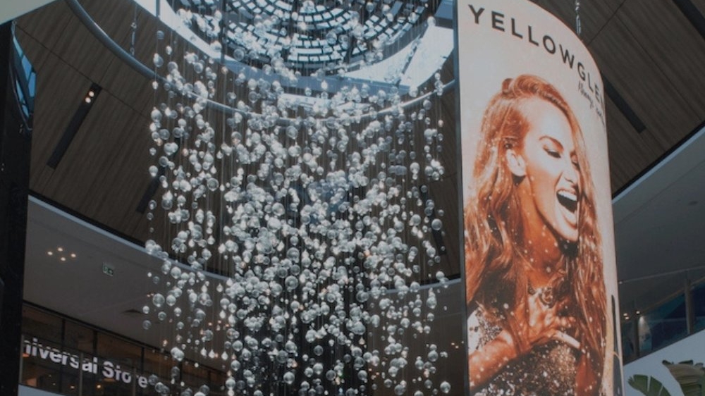 A large hanging flexible digital screen hangs in a public area showing an ad with a woman on it.